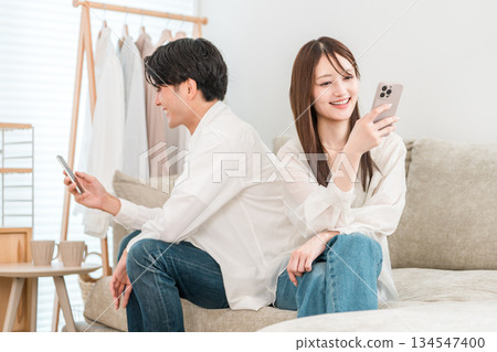 A young Asian couple sitting on the sofa in the living room at home and looking at their smartphone (misunderstanding, smartphone addiction) A young Asian couple sitting on the sofa in the living room at home and looking at their smartphone (misunderstanding, smartphone addiction) 134547400