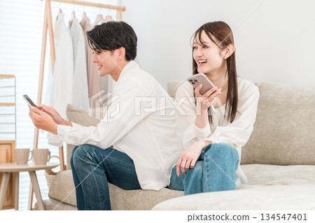 一對年輕的亞洲夫婦坐在家裡的客廳沙發上,一邊看著智慧型手機一邊聊天。 一對年輕的亞洲夫婦坐在家裡的客廳沙發上,一邊看著智慧型手機一邊聊天。 134547401