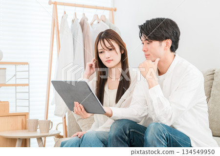 一對年輕的亞洲夫婦坐在家裡的客廳沙發上看平板電腦。 134549950