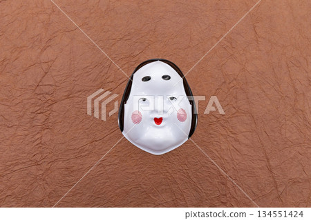 一張阿龜(禦福)面具置於棕色和紙背景的中央。這是節分/幸運符的圖案。 一張阿龜(禦福)面具置於棕色和紙背景的中央。這是節分/幸運符的圖案。 134551424