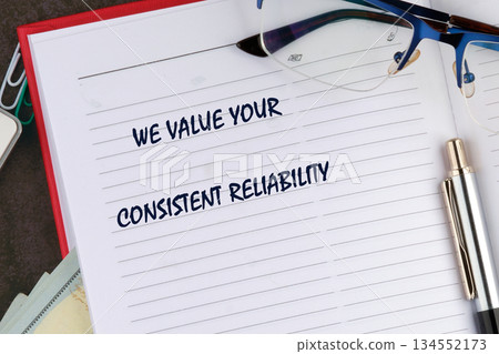 A lined notebook rests on a desk with the words WE VALUE YOUR CONSISTENT RELIABILITY written. A pen and glasses lay nearby, suggesting thoughtful planning and attention to detail A lined notebook rests on a desk with the words WE VALUE YOUR CONSISTENT RELIABILITY written. A pen and glasses lay nearby, suggesting thoughtful planning and attention to detail 134552173