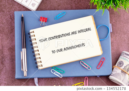 A notepad displays the text YOUR CONTRIBUTIONS ARE INTEGRAL TO OUR ACHIEVEMENTS. It sits on a blue notebook with a pen, clips, and money nearby, symbolizing gratitude 134552178