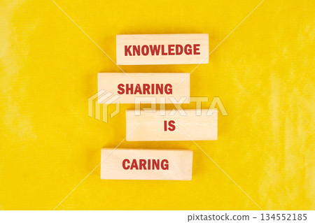 Four rectangular wooden blocks are stacked vertically, forming the phrase Knowledge Sharing is Caring. The red lettering adds a splash of color against a vibrant yellow backdrop 134552185