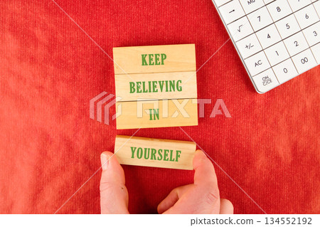 Hand positions a wooden block that reads YOURSELF to finish the phrase KEEP BELIEVING IN beside a calculator on red background, forming message of empowerment 134552192