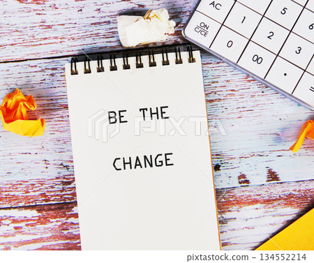 A notepad displaying be the change sits beside a calculator and crumpled paper on a white and brown weathered wooden surface, hinting at planning and transformation 134552214