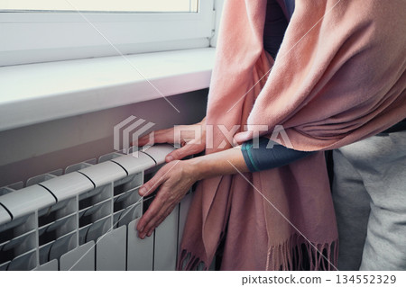 Woman warming hands on white radiator near window, feeling cold in her home 134552329