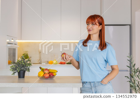 Young woman at kitchen bar takes apple from fruit bowl tosses it up bites and walks away happy smile 134552970