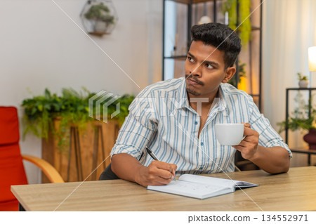 Indian man writes goals in notebook sips coffee ticks boxes smiles and plans day focused at desk 134552971