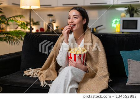 Young woman in blanket with popcorn laughs at movie or online show comments reacts cheerful evening Young woman in blanket with popcorn laughs at movie or online show comments reacts cheerful evening 134552982