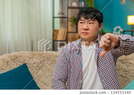 Asian man showing thumbs down, unhappy dislike expression signaling strong negative disagreement 134553340