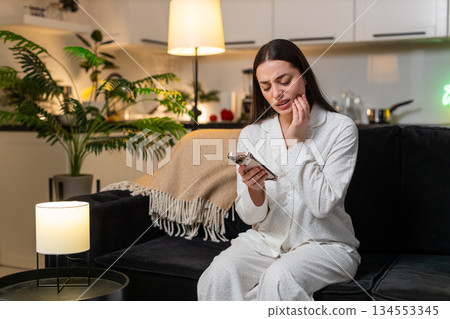 Young woman holds cheek tooth pain searches dentist on smartphone books visit dental care at night 134553345