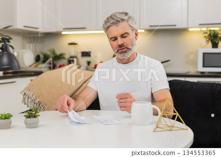 Middle-aged man sorts receipts on sofa makes paid due stacks organizes bills and keeps clear order 134553365