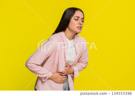 Young woman bending forward rubbing stomach showing painful expression suffering from abdominal ache Young woman bending forward rubbing stomach showing painful expression suffering from abdominal ache 134553396
