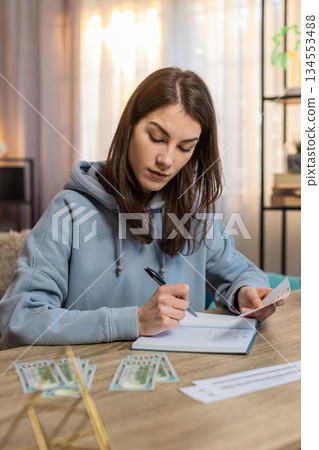 Young woman sitting at table looking at receipts and counting money while making shopping list 134553488
