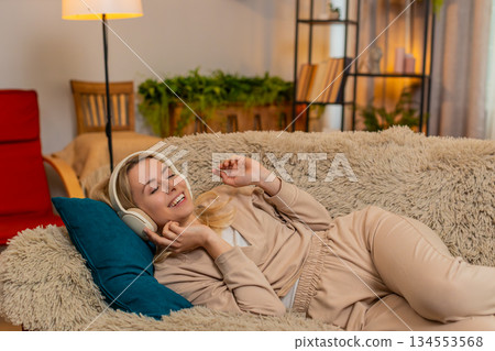 Woman lying on sofa with eyes closed, listening to music through headphones in peaceful relaxation 134553568