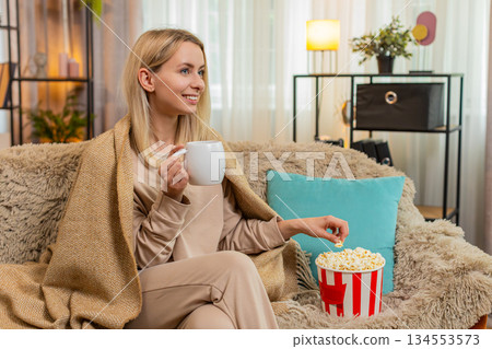 Adult woman wrapped in blanket sitting on sofa with cup of tea watching series thoughtfully at home Adult woman wrapped in blanket sitting on sofa with cup of tea watching series thoughtfully at home 134553573