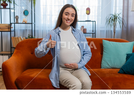 Happy pregnant woman stroking big belly with love and showing thumbs up sign sitting on home sofa Happy pregnant woman stroking big belly with love and showing thumbs up sign sitting on home sofa 134553589