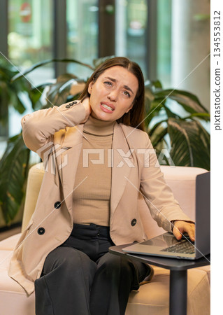 Tired young businesswoman suffers from spine neck muscle pain working on laptop in office lobby Tired young businesswoman suffers from spine neck muscle pain working on laptop in office lobby 134553812
