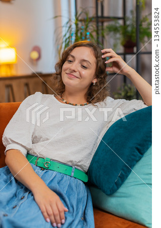 Happy carefree young Caucasian woman sitting comfortably on sofa thinking in living room at home Happy carefree young Caucasian woman sitting comfortably on sofa thinking in living room at home 134553824