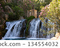 Cascading waterfall flows over rocky terrain surrounded by vibrant greenery and blooming flowers, creating a serene natural landscape with tranquil atmosphere 134554032