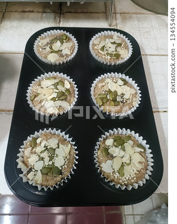 Homemade muffin batter in baking tray with seed topping, almond slice, paper cups, rustic kitchen background Homemade muffin batter in baking tray with seed topping, almond slice, paper cups, rustic kitchen background 134554094