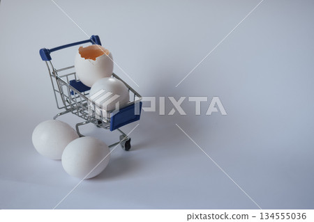Broken egg in a shopping cart on white, symbolizing economic fragility and rising food prices 134555036