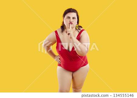 Portrait of overweight woman wearing swimsuit shows gesture of silence on background in studio. Portrait of overweight woman wearing swimsuit shows gesture of silence on background in studio. 134555146