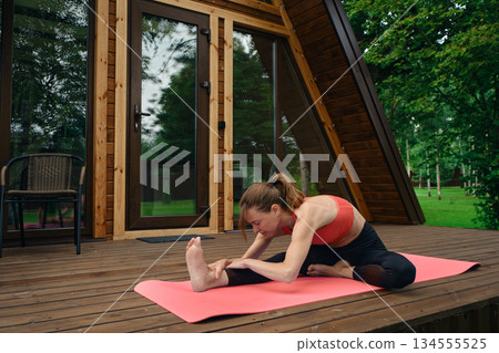 Woman practices yoga on a pink mat outside a wooden cabin in the morning 134555525