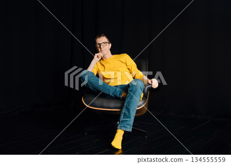Middle-aged in yellow shirt sitting in a chair with one leg crossed on a black background Middle-aged in yellow shirt sitting in a chair with one leg crossed on a black background 134555559