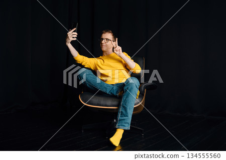 Middle-aged man sits on chair and takes selfie in dark room with yellow shirt and blue jeans Middle-aged man sits on chair and takes selfie in dark room with yellow shirt and blue jeans 134555560
