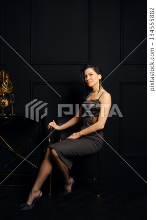 Woman seated on chair next to a table with a gold decoration in a dark room Woman seated on chair next to a table with a gold decoration in a dark room 134555882