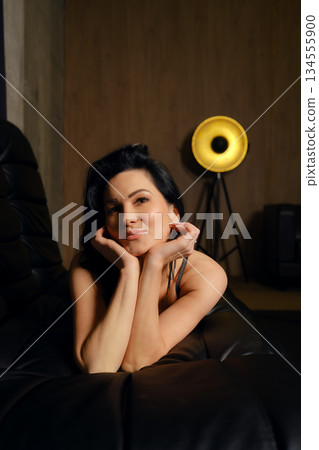 Woman poses with hands on chin in modern indoor space with unique lamp in background Woman poses with hands on chin in modern indoor space with unique lamp in background 134555900