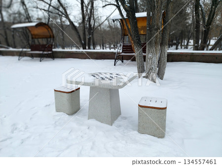 A concrete outdoor chessboard table with two matching stools, dusted with snow, sits in a snowy park setting with bare trees and blurred structures in the background 134557461