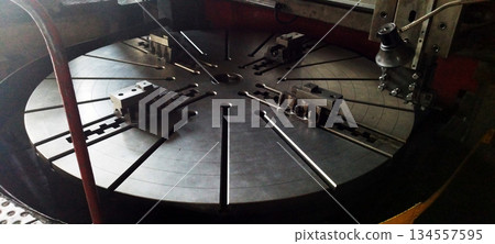 The massive, slotted rotating table of a heavy-duty vertical turning lathe, featuring several large metal chuck jaws, sits under dim industrial lighting 134557595