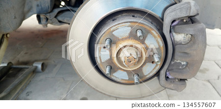 A close-up of a vehicle wheel hub assembly, showing a brake rotor, rusted lug studs, the brake caliper, and the center axle nut, likely undergoing maintenance A close-up of a vehicle wheel hub assembly, showing a brake rotor, rusted lug studs, the brake caliper, and the center axle nut, likely undergoing maintenance 134557607