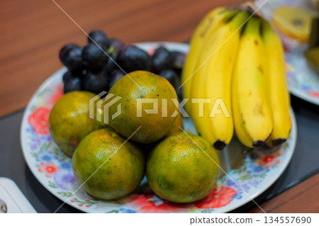 Close-up Studio Shot of Mixed Fruit Assortment Featuring Green Oranges, Yellow Bananas, and Dark Grapes on a Decorative Plate with Shallow Depth of Field Close-up Studio Shot of Mixed Fruit Assortment Featuring Green Oranges, Yellow Bananas, and Dark Grapes on a Decorative Plate with Shallow Depth of Field 134557690