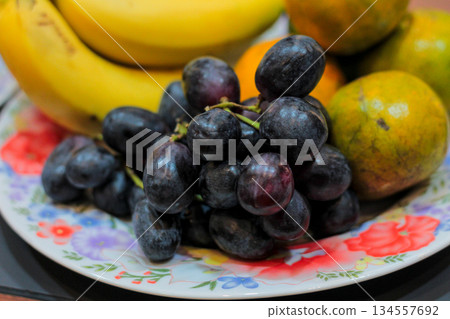 Close-up macro shot of ripe black grapes centered on a patterned floral dish, featuring soft focus on citrus fruits and bananas in the vibrant background. Close-up macro shot of ripe black grapes centered on a patterned floral dish, featuring soft focus on citrus fruits and bananas in the vibrant background. 134557692