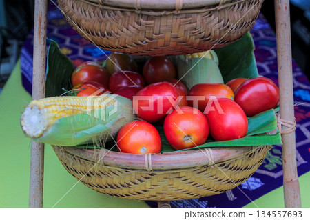 Close-up high-angle shot of vibrant red heirloom tomatoes and yellow corn displayed in rustic two-tiered bamboo storage basket Close-up high-angle shot of vibrant red heirloom tomatoes and yellow corn displayed in rustic two-tiered bamboo storage basket 134557693