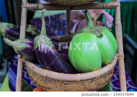 Macro shot capturing the texture and contrast of raw, organically grown Solanum melongena (purple eggplant) next to a round green Thai eggplant variety (Solanum xanthocarpum) in a rattan carrier. Macro shot capturing the texture and contrast of raw, organically grown Solanum melongena (purple eggplant) next to a round green Thai eggplant variety (Solanum xanthocarpum) in a rattan carrier. 134557694