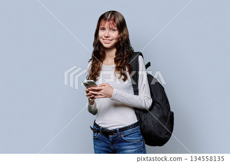 Portrait of beautiful girl teenage student with backpack, smartphone on gray background 134558135