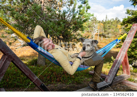 Woman relaxing in hammock with dog wearing outdoor clothing in forest nature Woman relaxing in hammock with dog wearing outdoor clothing in forest nature 134562410