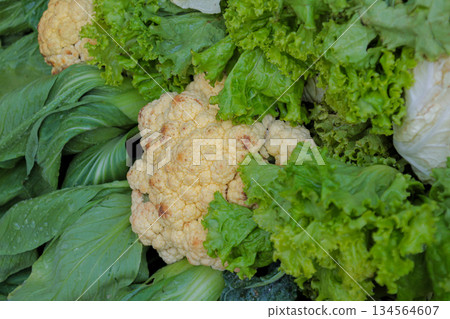 Close-up view of fresh cauliflower head surrounded by assorted green lettuce and leafy vegetables at market stall. Close-up view of fresh cauliflower head surrounded by assorted green lettuce and leafy vegetables at market stall. 134564607