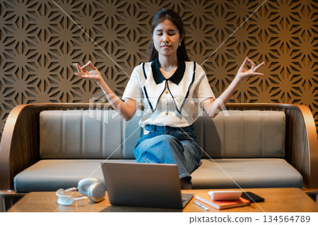 A young woman practices meditation in a stylish office lounge, sitting on a sofa with a laptop and headphones nearby. A young woman practices meditation in a stylish office lounge, sitting on a sofa with a laptop and headphones nearby. 134564789