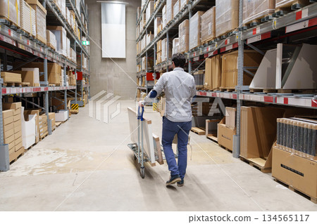 Man walking through furniture warehouse with cart and boxed furniture 134565117