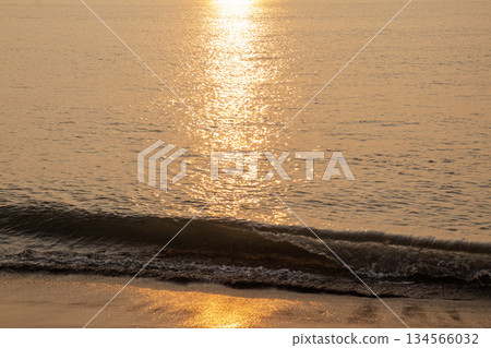 Golden sunlight path reflecting brightly on the ocean water surface with a small wave breaking onto the sandy beach during sunset. Golden sunlight path reflecting brightly on the ocean water surface with a small wave breaking onto the sandy beach during sunset. 134566032