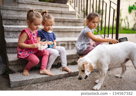 Three girls enjoy snack time with their playful dog outdoors 134567382