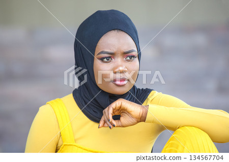 african woman in Black Hijab and Yellow Outfit Sitting Thoughtfully Against a Neutral Background african woman in Black Hijab and Yellow Outfit Sitting Thoughtfully Against a Neutral Background 134567704