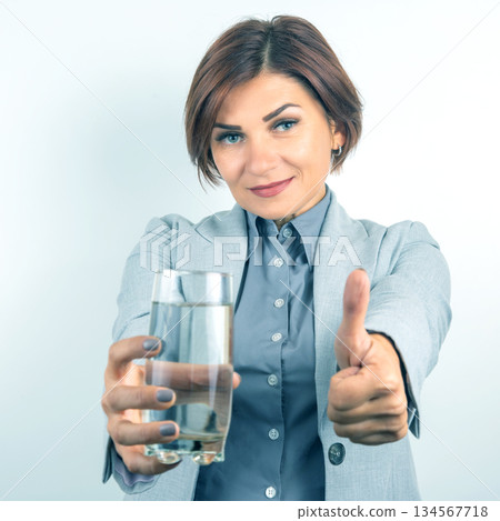 Professional woman smiling and showing a glass of water with a thumbs up for hydration benefits Professional woman smiling and showing a glass of water with a thumbs up for hydration benefits 134567718