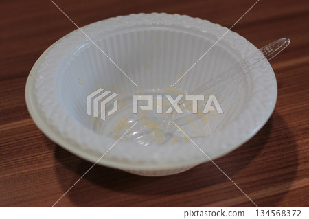 White disposable plastic bowl with residual food and clear plastic spoon resting on brown wooden table after finishing meal. 134568372