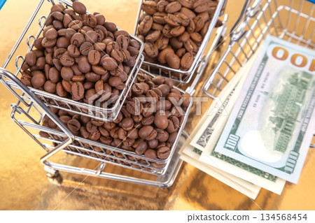 A stack of 100 bills lies beneath a cart of coffee beans on a golden backdrop 134568454